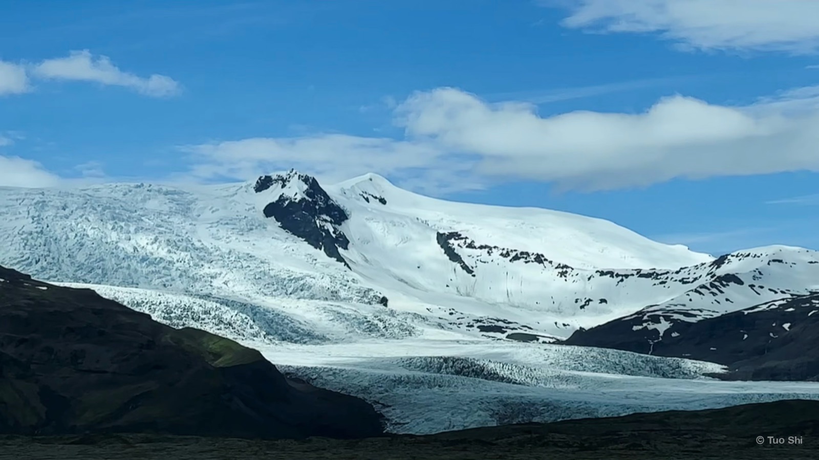 A white glacier flowing down between dark mountains under a blue sky