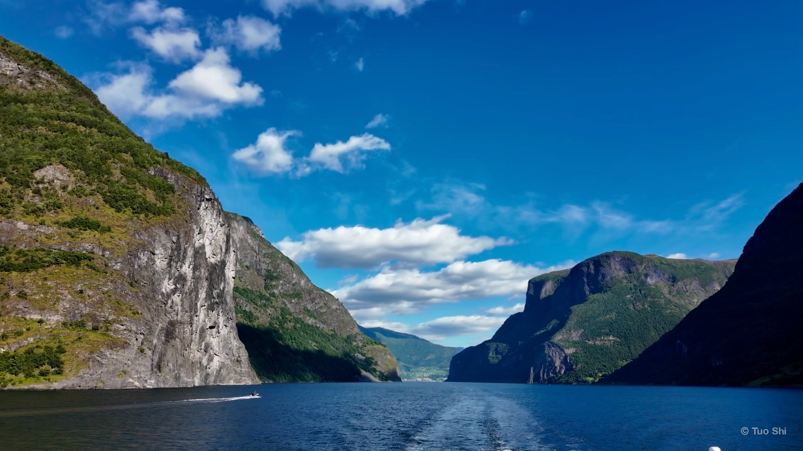 A ferry wake leading into a deep blue fjord between steep mountain walls