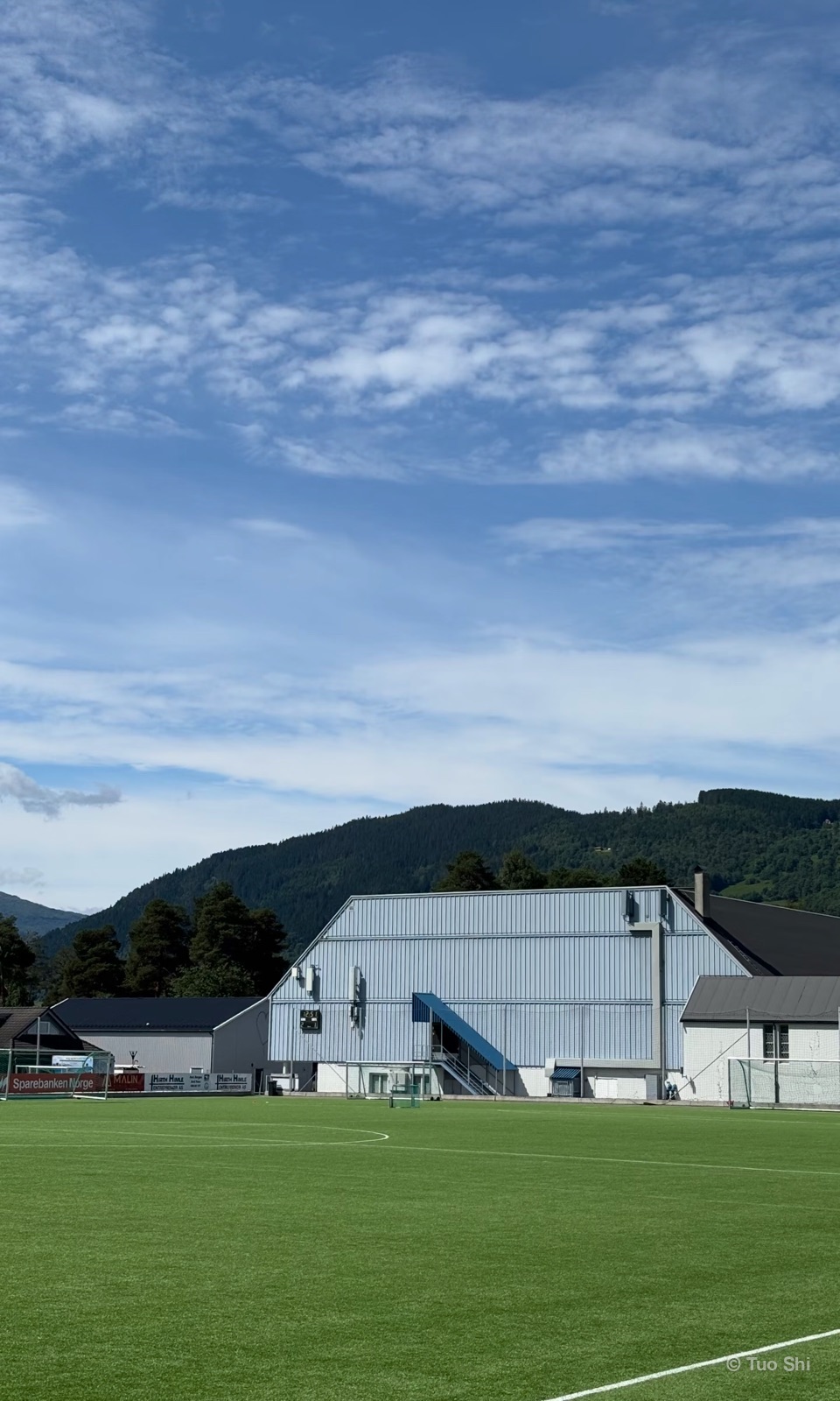 A football pitch and sports hall beneath forested hills and a wide sky