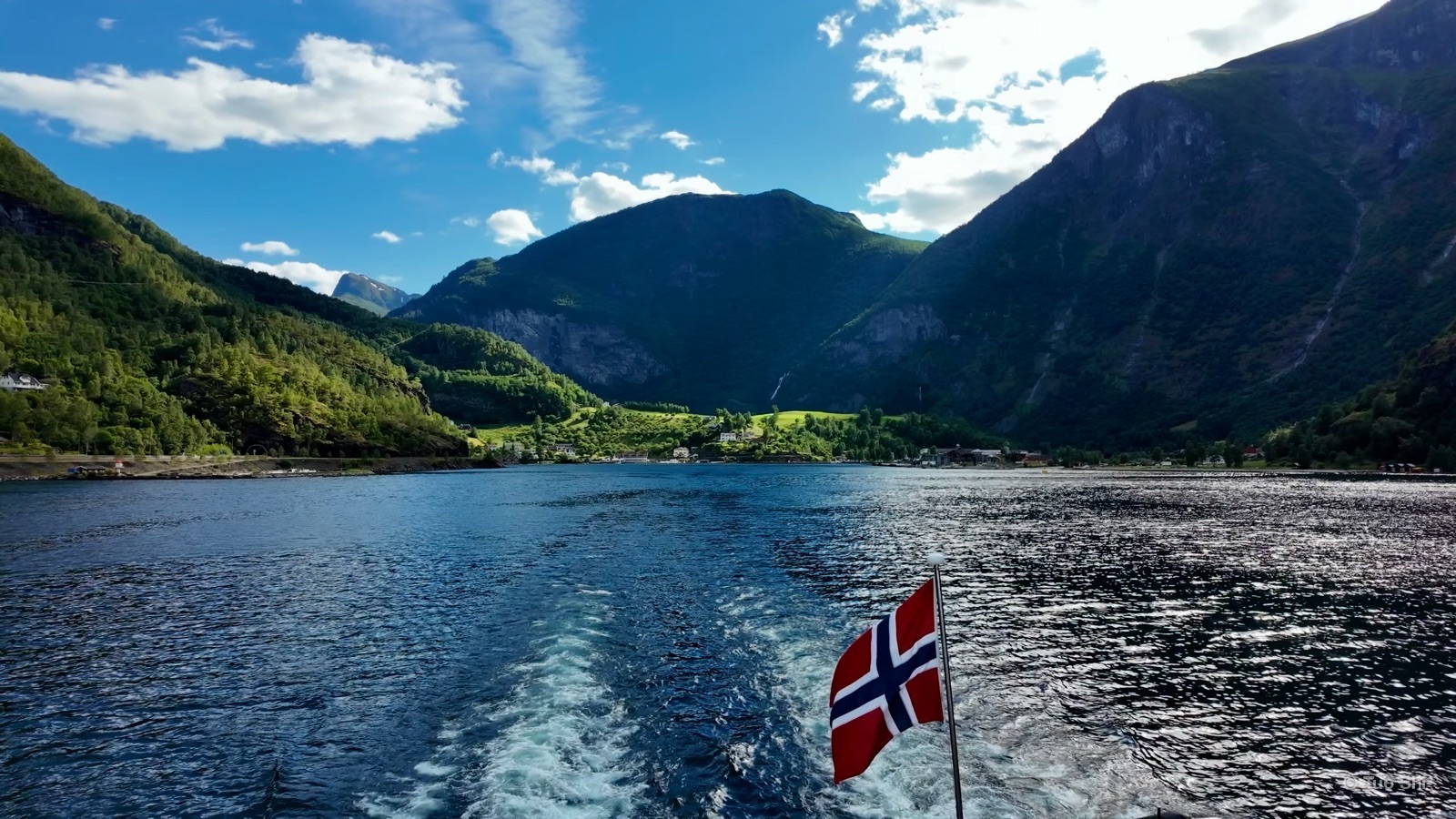The Norwegian flag flying over a boat's wake on a sunlit fjord