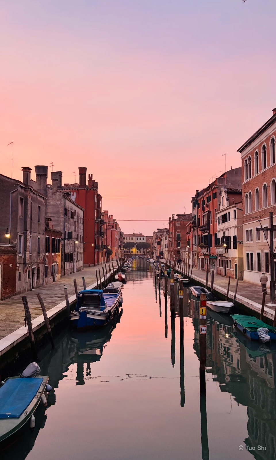 A calm Venetian canal lined with boats and houses in pink evening light